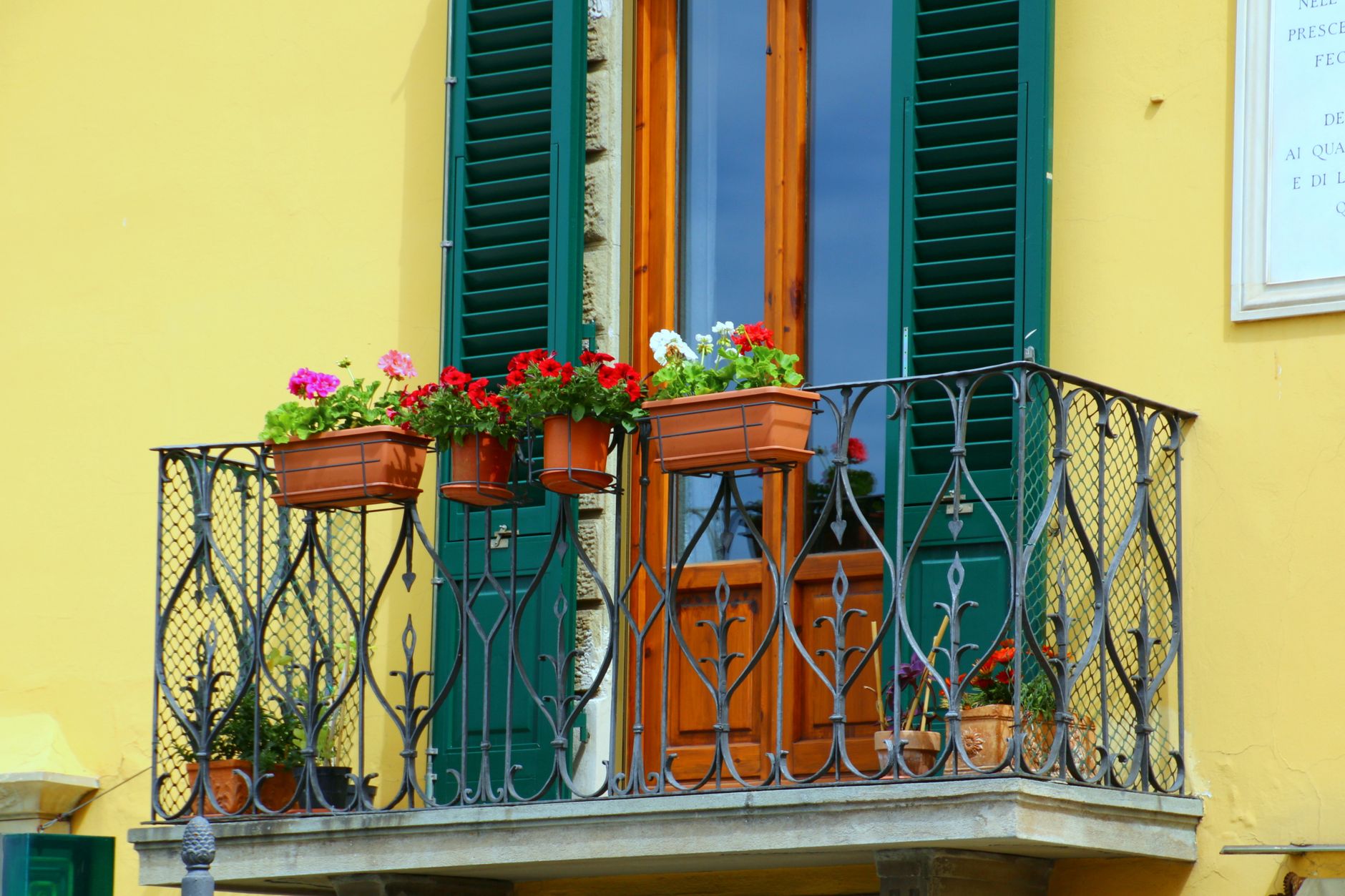 Colorful flowers adorn a quaint balcony with green shutters and a yellow wall.