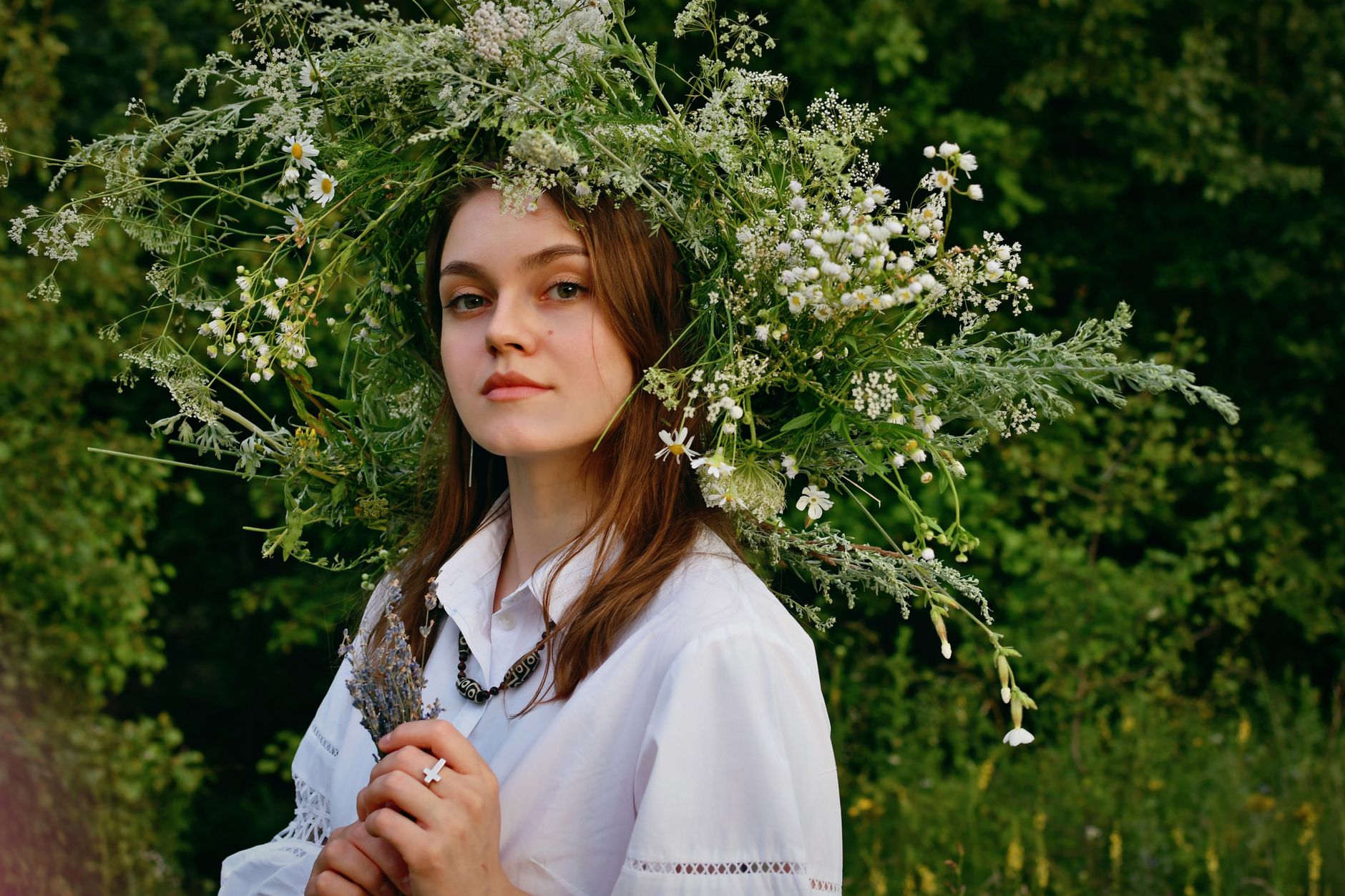 Portrait of a woman with a floral wreath standing outdoors against lush greenery.